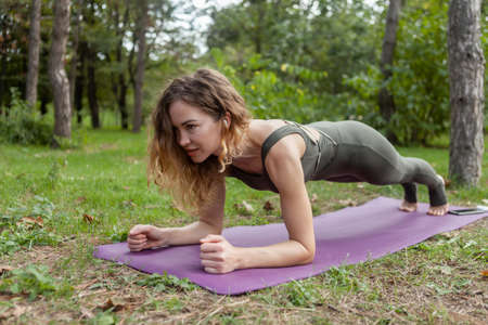 Attractive yogi woman with curly hair is practicing plank exercise while training endurance in forestの写真素材