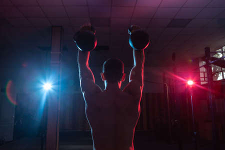 Muscular Caucasian man with naked torso training overhead with kettlebells in red blue neon gradient light.の写真素材