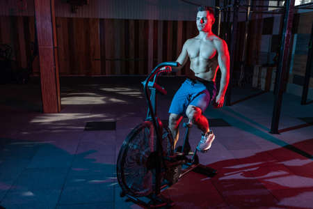 Muscular man training on air bike in red-blue neon light. Cross training