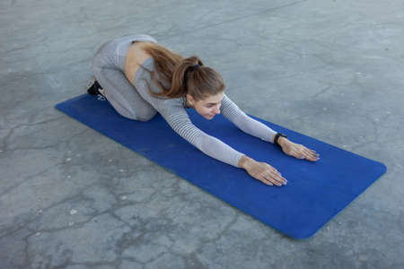 Young caucasian blond fitness woman is exercising stretching while sitting on a mat on a concrete floor. healthy lifestyleの写真素材