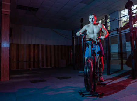 Muscular man training on air bike in red-blue neon light. Cross training