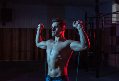 Muscular powerful man with naked torso trains with expander (fitness rubber bands) in red blue neon...