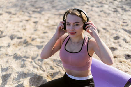 Attractive fit woman listening to music with headphones while sitting on mat on the beach on a bright sunny dayの写真素材