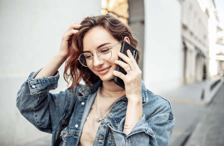 Portrait of a charismatic stylish and modern woman in trendy clothes with glasses talking on the phone. Attractive young woman walking along the urban streetの写真素材