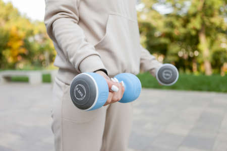 Young sportive brunette woman in sportswear trains muscles with dumbbells outdoors. healthy...