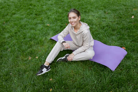 Happy fit woman has rest time while sitting on the mat in the park. Fitness or yoga outdoors