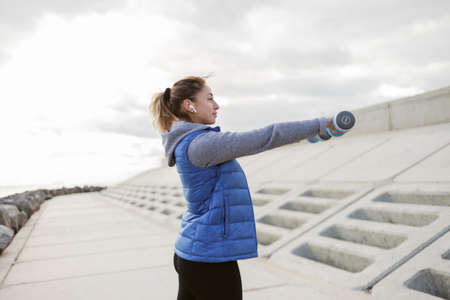 Attractive fit woman in warm sportswear trains shoulder muscles with dumbbells outdoors. healthy...