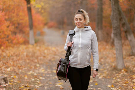 Attractive sporty woman with bag goes to workout in autumn forest or park