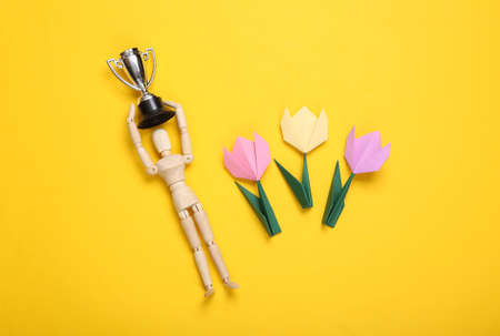 Origami tulips and wooden puppet holding the winner's cup on a yellow background. Victory concept. flat lay. top viewの写真素材