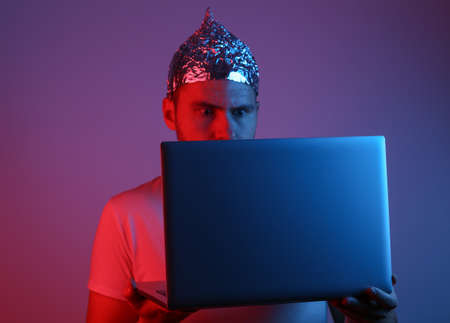 Crazy Bearded Man in a foil hat with a laptop in red blue neon light. Conspiracy