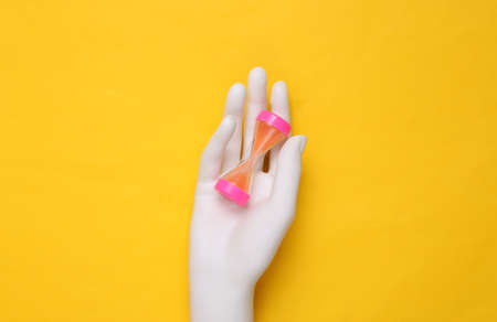Mannequin hand holding hourglass on yellow background. top view. minimalism