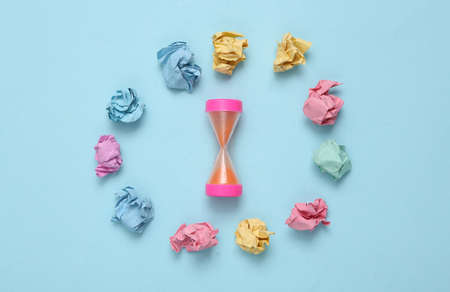 Hourglass and crumpled balls of colored paper on a blue background. Lack of ideas conceptの写真素材