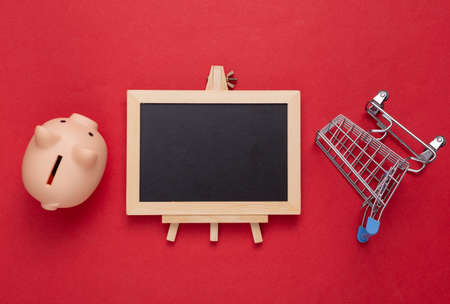 Shopping cart and piggy bank, empty chalk board on a red background. copy spaceの写真素材