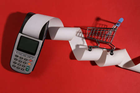 Payment terminal with cash tape and mini shopping trolley on red bright background. top view