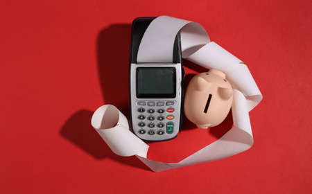 Payment terminal with cash tape and piggy bank on red bright background. Save money concept. top...