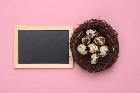 Easter composition. Quail eggs in a nest and empty chalk board with copy space on a pink pastel...