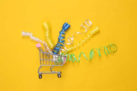 Colored streamer in shopping trolley on yellow background. Holiday shopping concept
