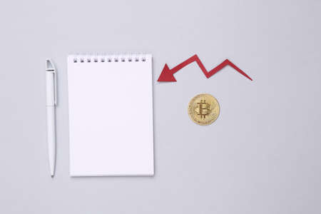 Notepad and golden bitcoin with falling arrow on gray background. Business and finance, trading. bitcoin price drop conceptの写真素材