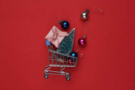 Mini shopping cart with christmas balls, mini christmas tree and gift box on red backgroundの写真素材