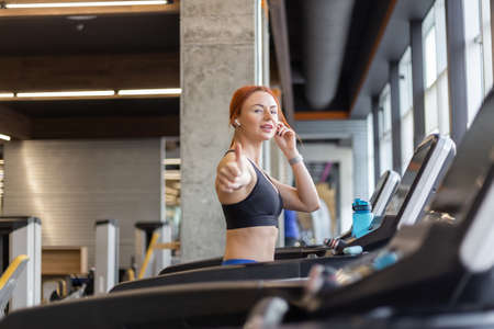 Red-haired fitness woman showing thumbs up in modern gym. Happy fit slim woman works out on a...