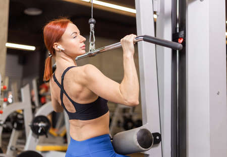 A red-haired fit woman in sportswear trains back muscles in a lat exercise machine. Young fitness...