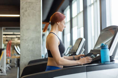 Portrait of a red-haired fitness woman in a modern gym. Fit slim woman works out on a treadmill,...