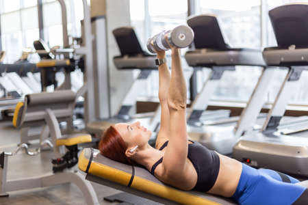 Athletic red-haired woman trains chest muscles with dumbbells in her hands while lying on an incline...