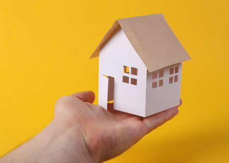 Hand holds mini paper home figurine on yellow background. Protection concept, securityの写真素材
