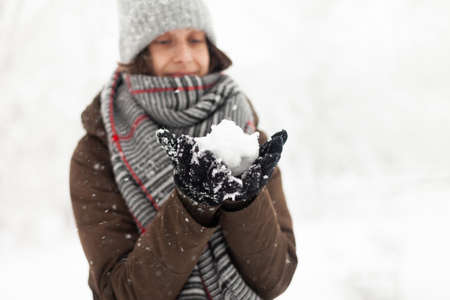 Cheerful woman in warm clothes holds snow in her hands in winter snowy weather.