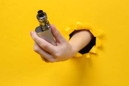 Female hand holds vaping device through torn hole yellow paper. concept art.