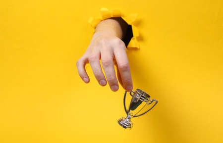Female hand holds winner cup through torn hole yellow paper. concept art. minimalismの写真素材
