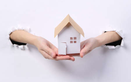 Female hands holding house figure through torn hole white paper.の写真素材