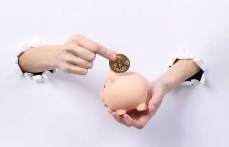 Female hands put bitcoin in piggy bank through torn hole white paper. concept art