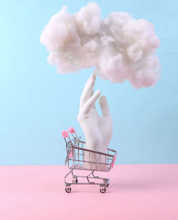 Minimalistic composition with hand in shopping trolley and fluffy cloud on a blue pink pastel...