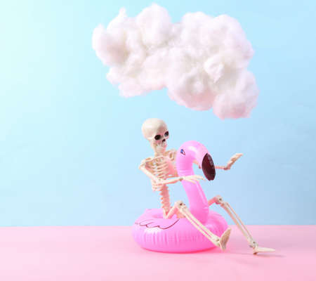 Skeleton with inflatable flamingo and floating fluffy cloud on blue pink pastel background. concept art. minimalist ideasの写真素材