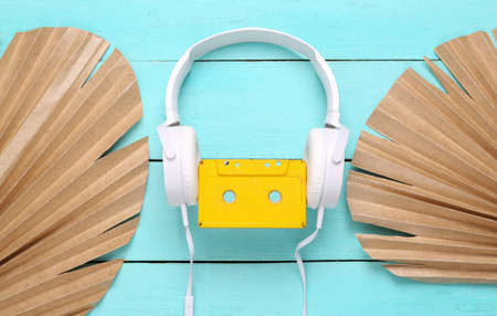 Stereo headphones with audio cassette and dry palm leaves on blue wooden background. top view....