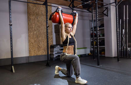 Athletic strong woman exercising with heavy functional training bag in modern gym