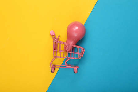 Mini pink supermarket trolley with light bulb on a blue-yellow background. fresh ideas. creative layout. top view. flat layの写真素材