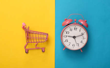 Mini pink supermarket trolley with alarm clock on blue-yellow background. creative layout. top view....