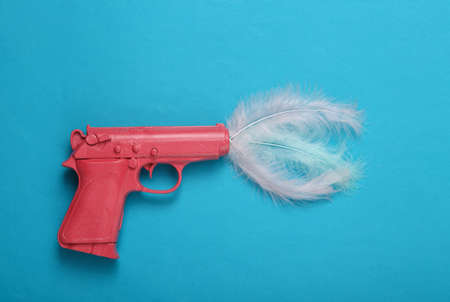Pink pistol gun with feathers on blue background. creative concept. Minimalist flat lay. Minimal layout.Top viewの写真素材
