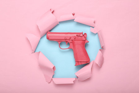 Pink pistol gun on a blue-pink torn background. creative concept. Minimalist flat lay. Minimal...