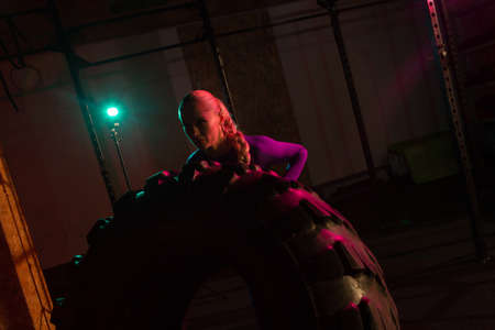Fit female athlete flipping huge tire. Woman lifts a heavy wheel in pink green neon light. Muscular...