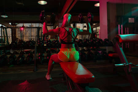 Fit woman in sportswear working out with heavy dumbbells while sitting on bench in pink green neon lightの写真素材