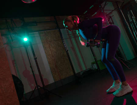 Young fit slim woman working out with dumbbells in her hands in pink green neon gradient light at...