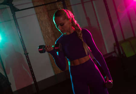 Young fit slim woman trains biceps with dumbbells in her hands in pink green neon gradient light at gym. Sport, fitness, lifestyle conceptの写真素材