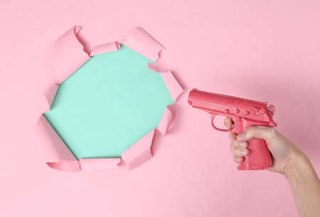 Hand holds pink pistol on blue-pink paper background with ripped space for copy space. Concept art,...