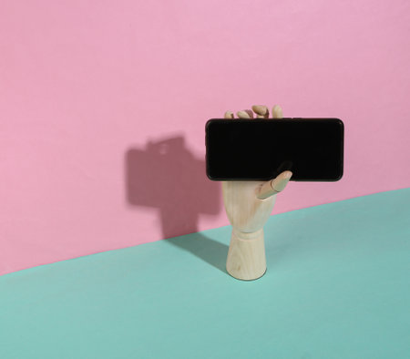 Wooden hand holding smartphone on blue pink pastel background. trendy shadow. concept art....