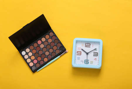 Alarm clock and eye shadow palette on yellow background. makeup time. top view