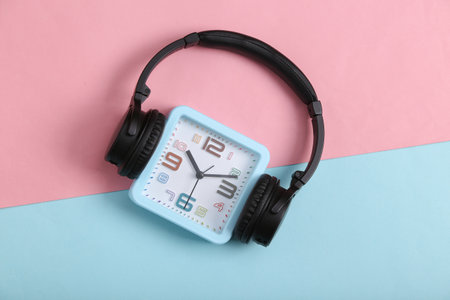 Square alarm clock with headphones on pink blue background. music time. top view