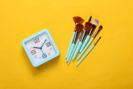 Alarm clock and makeup brushes on yellow background. makeup time. top view
